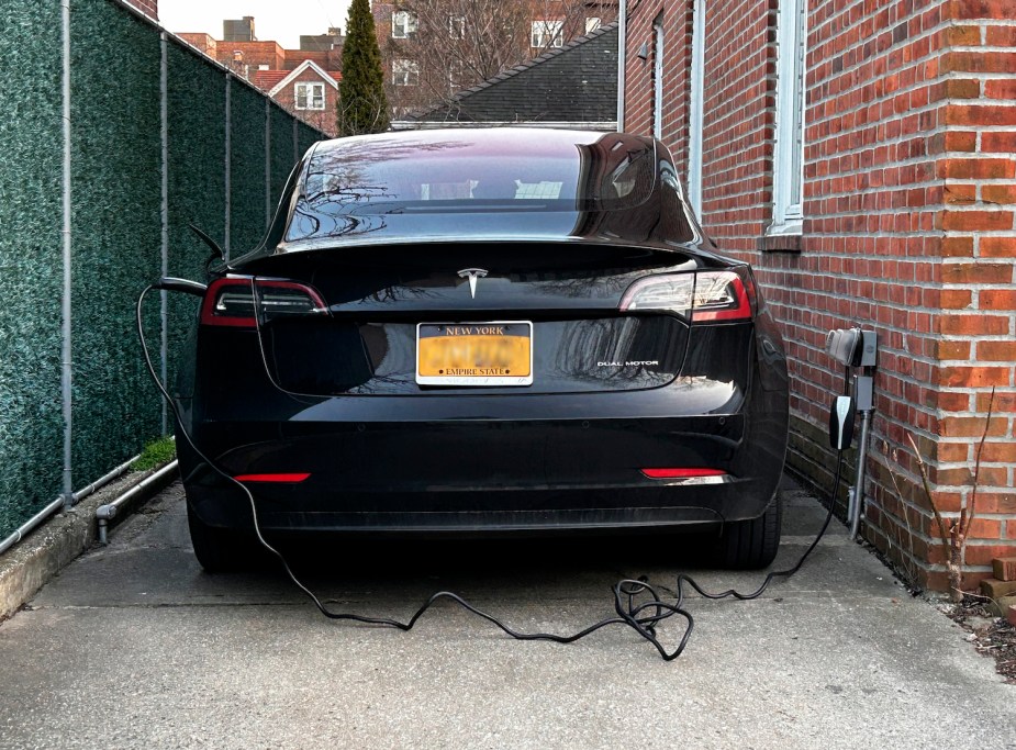 A Tesla electric vehicle charging in a driveway, city buildings visible in the background.