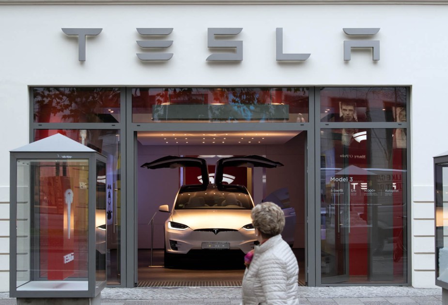 A white Model Y SUV parked in a Tesla showroom, a passerby in the foreground looking at the electric vehicle.