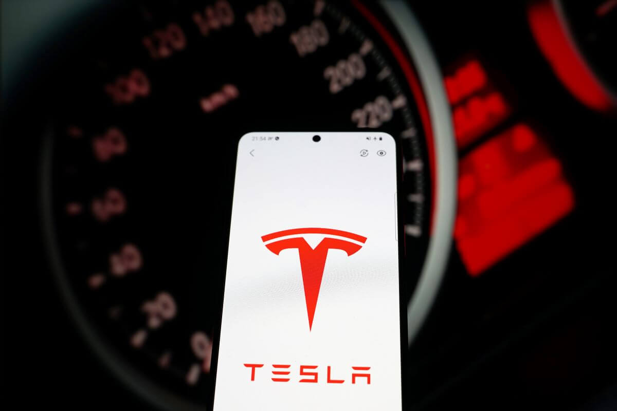 The Tesla app, signified by the automaker's logo on a smartphone screen, with a car dashboard behind it