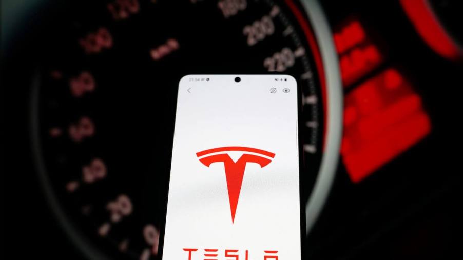 The Tesla app, signified by the automaker's logo on a smartphone screen, with a car dashboard behind it