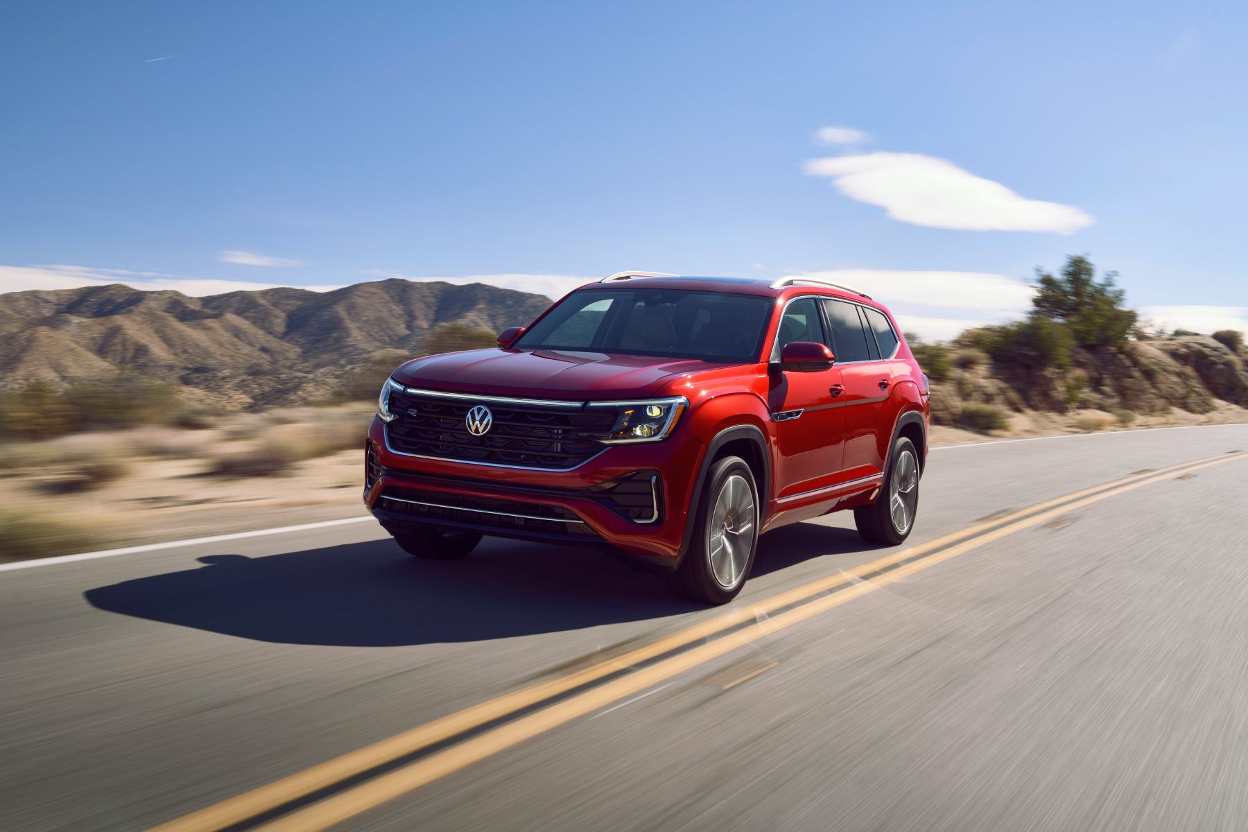 A red 2024 Volkswagen Atlas drives down a road with mountains in the background.