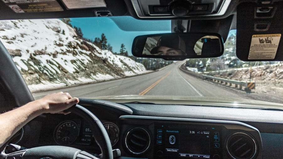A man's hand on the steering wheel of his Toyota Tacoma pickup truck, snowy mountains visible through the windshield.