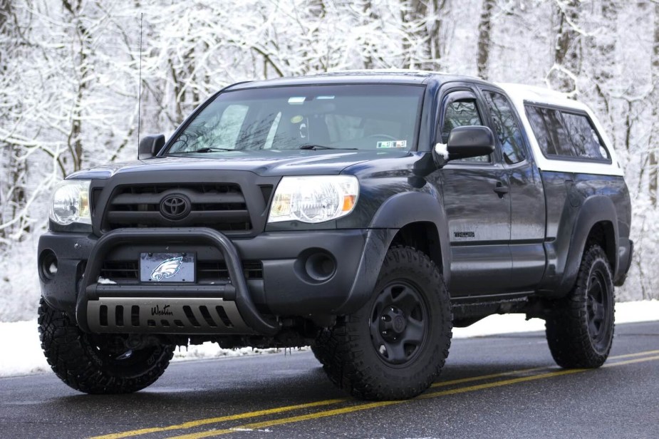Black Toyota Tacoma with a white cap is parked on a paved road with snow-covered trees visible in the background.