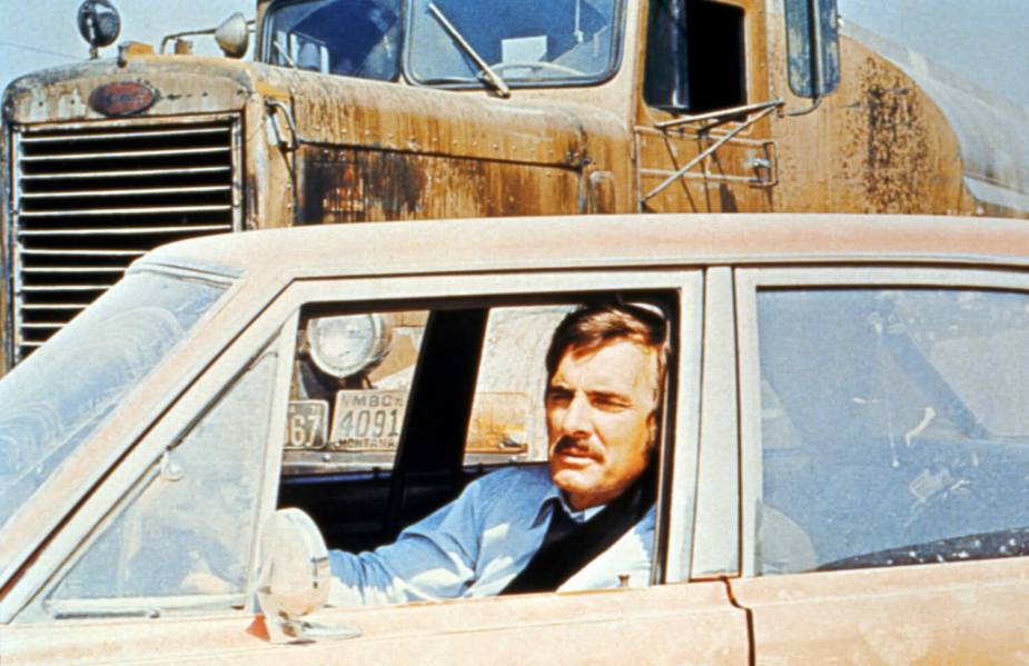 Dennis Weaver sits in the driver's seat of a Plymouth Valiant sedan on the set of Duel, a Peterbilt 281 seen in the background.