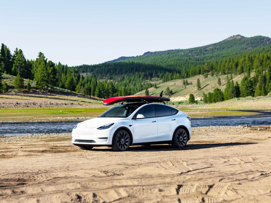 A white Tesla Model Y with a red paddleboard on its roof in front of a river and mountains.