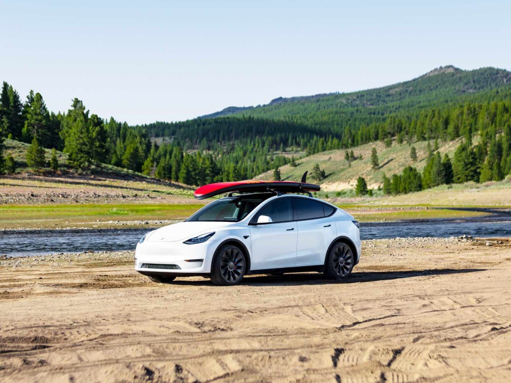 A white Tesla Model Y with a red paddleboard on its roof in front of a river and mountains. 