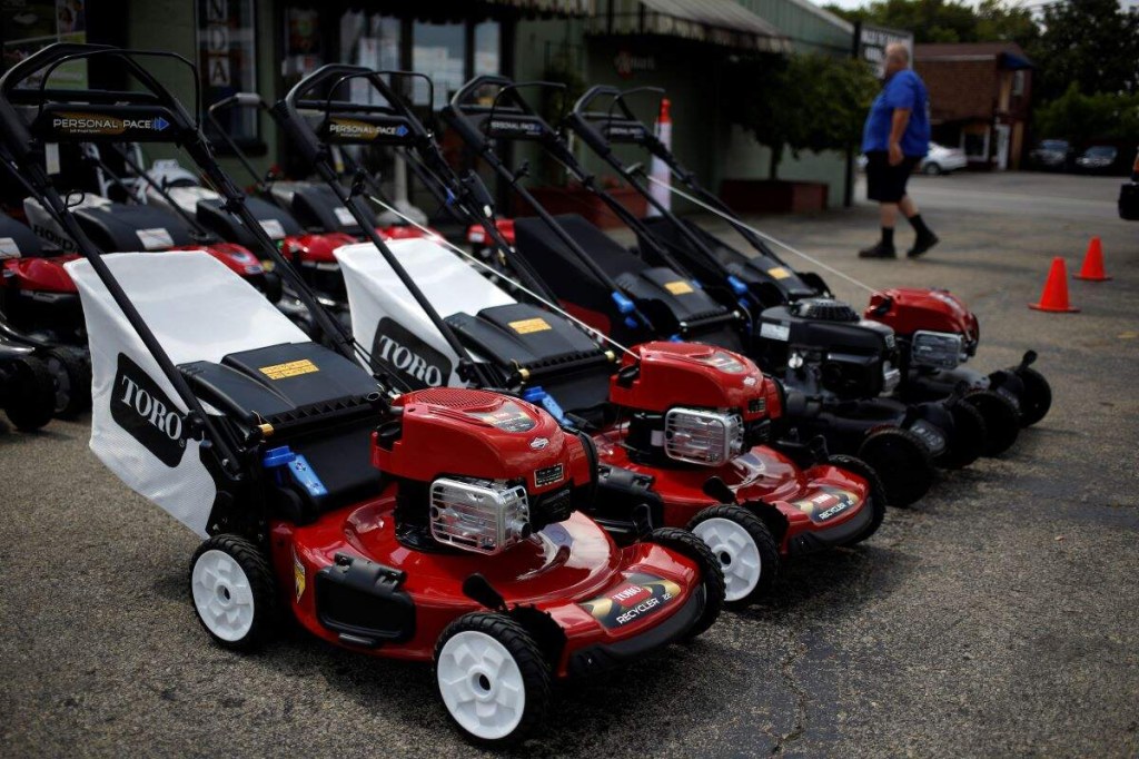 A row of lawn mowers, that may help with the consideration of electric vs. gas lawn mowers, all lined up outside of building. 