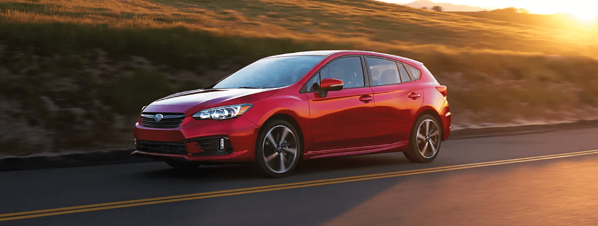 A red 2023 Subaru Impreza Hatchback model driving on a country highway as the sun sets behind it