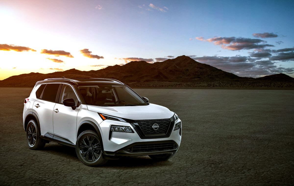 A white 2023 Nissan Rogue compact SUV model parked on dry and empty terrain in front of a sunset above a mountain.