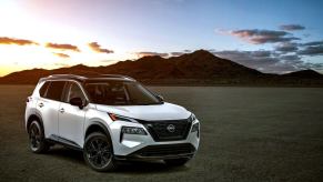 A white 2023 Nissan Rogue compact SUV model parked on dry and empty terrain in front of a sunset above a mountain
