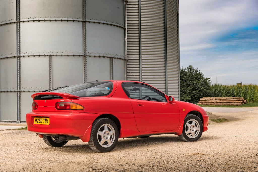 A red 1994 Mazda MX-3 parked next to a grain silo