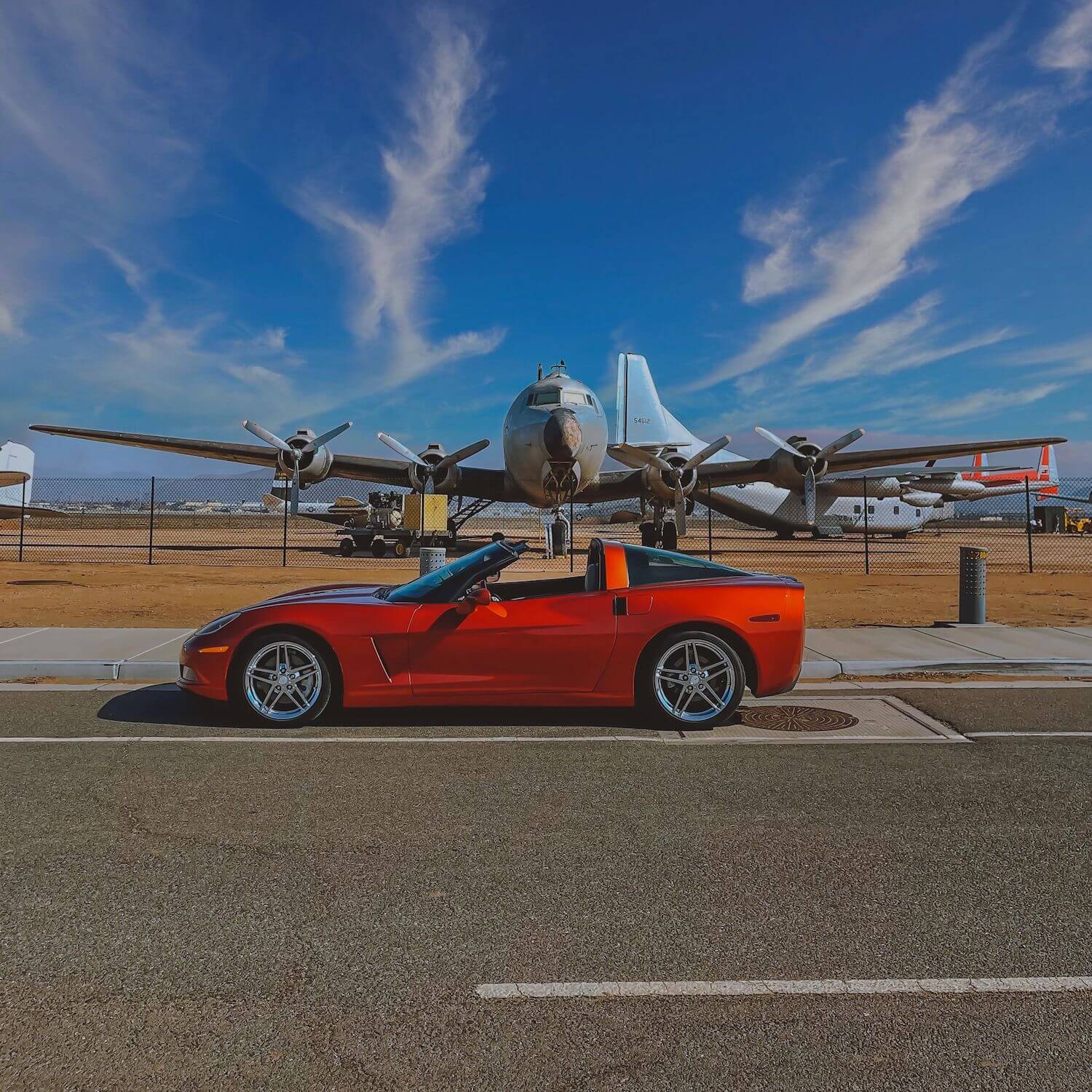 A burnt orange Corvette parked in front of an airport, planes visible in the background.