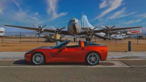 A burnt orange Corvette parked in front of an airport, planes visible in the background.