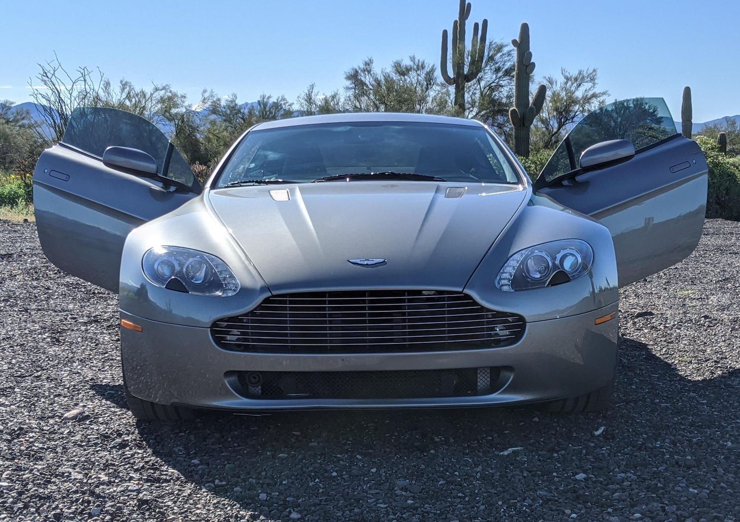 The grille of a used silver Aston Martin Vantage supercar with its doors open, cactuses visible in the background.