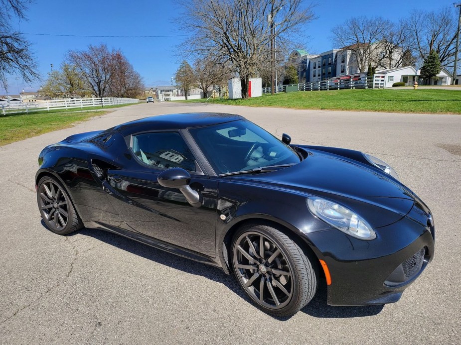 A used black Alfa Romeo 4C supercar in a parking lot, a farm with white picket fences visible in the background.