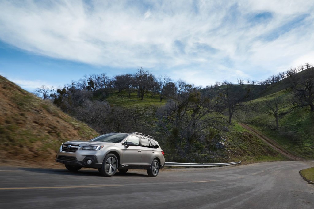 A silver Subaru Outback drives up an inclined road with a hilly terrain in the background. 