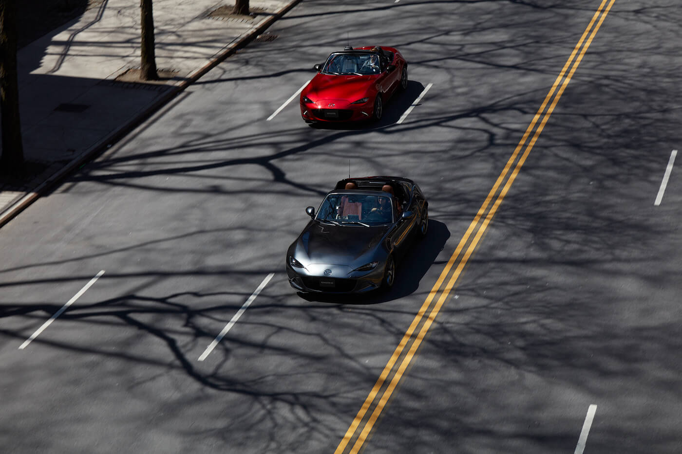 Two Mazda MX-5 Miata (one gray, the other black) models on a city street.