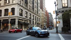 A gray Mazda MX-5 Miata turning on a city street in front of some buildings