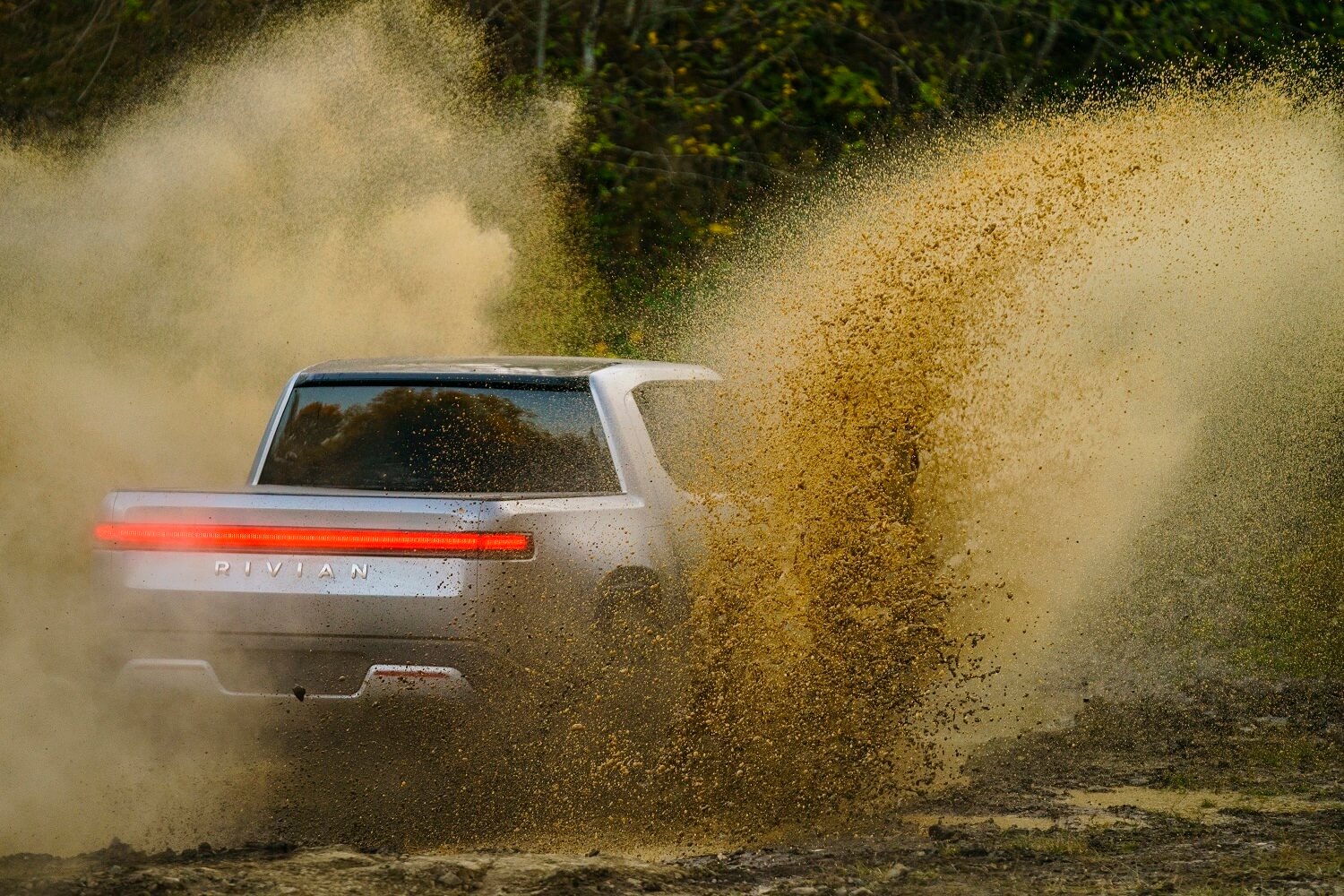A Rivian R1T electric truck racing across a dirt road, dust visible in the foreground.