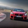 A red 2023 BMW M2 speeding down a track with storm clouds in the background.