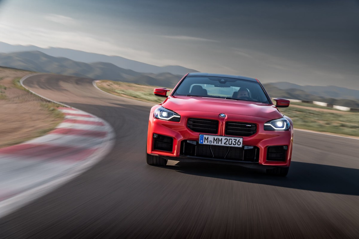 A red 2023 BMW M2 speeding down a track with storm clouds in the background.