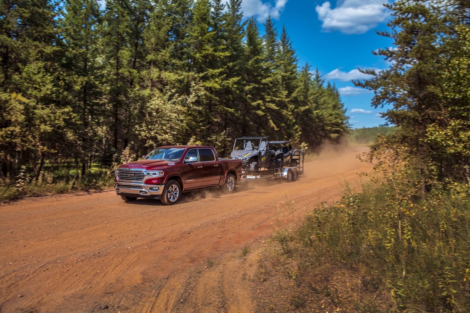 A red 5th-gen Ram 1500 pickup truck pulls a trailer down a dirt road, pin trees visible in the background.