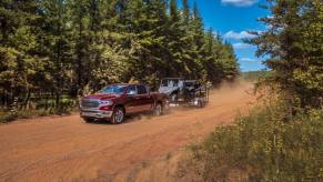A red 5th-gen Ram 1500 pickup truck pulls a trailer down a dirt road, pin trees visible in the background.