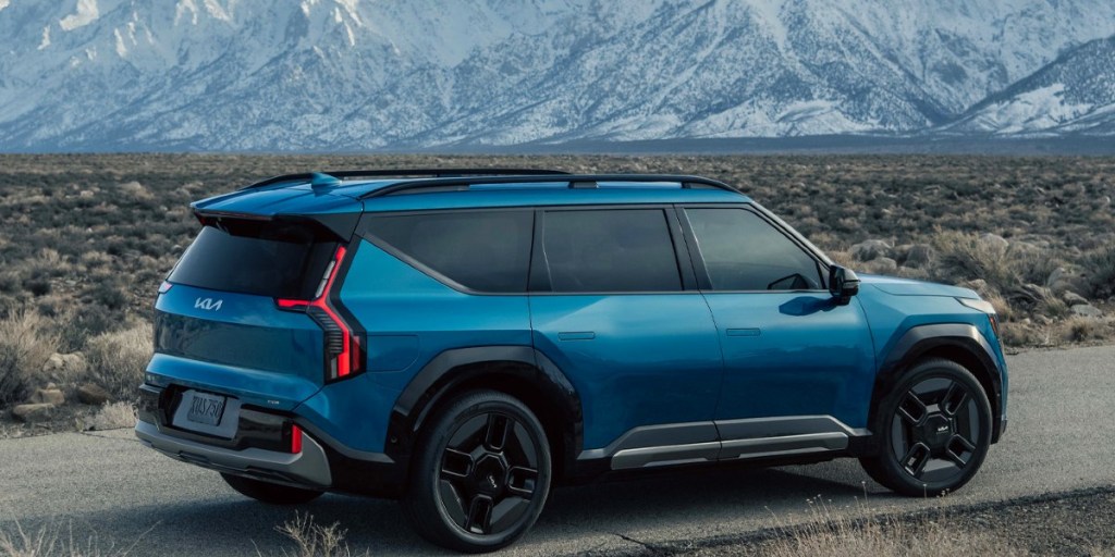 A blue 2024 Kia EV9 midsize electric SUV is driving on the road (side profile).