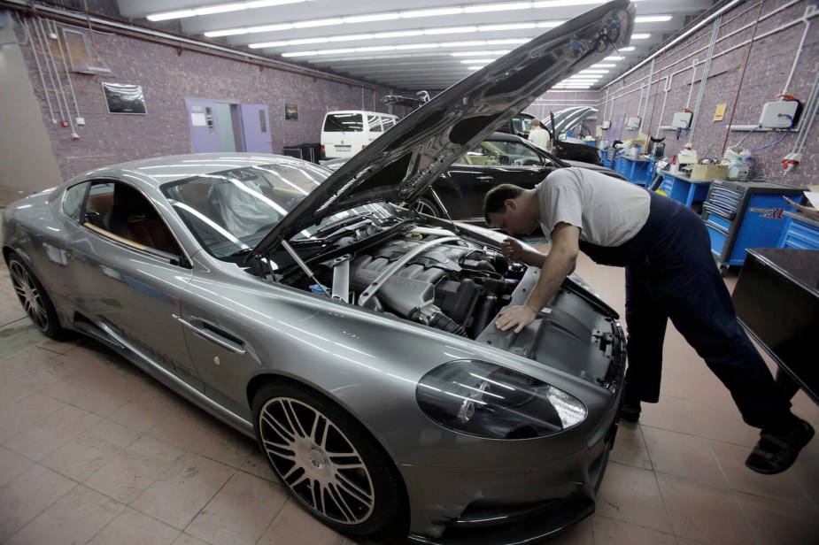 A mechanic leans over the hood of a used Aston Martin DB9 supercar, a crowded auto shop visible in the background.
