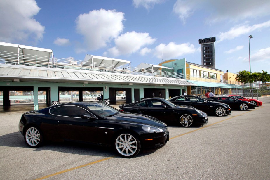 A lineup of supercars including an Aston Martin DB9 parked by a race track.