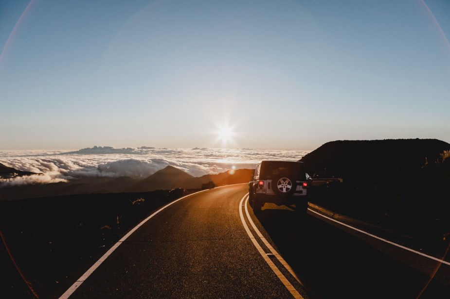 A Jeep Wrangler on a road trip, navigating a two-lane road over a mountain pass, the sunset in the background.