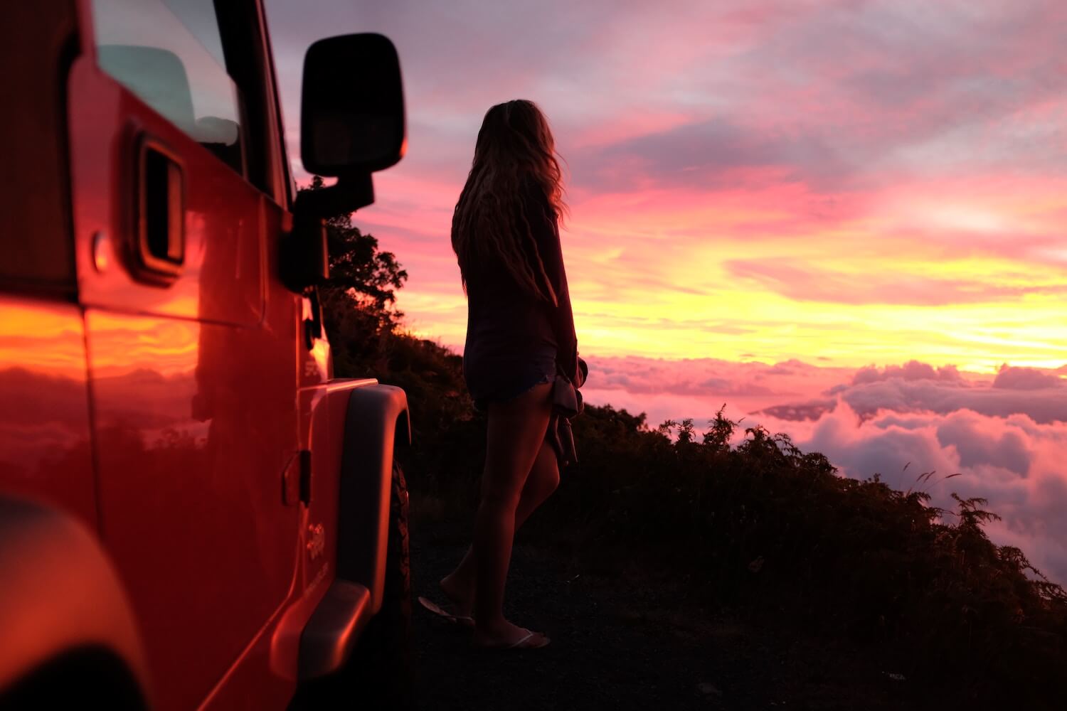 A woman stands next to a Jeep Wrangler, watching the sun set from atop a mountain.