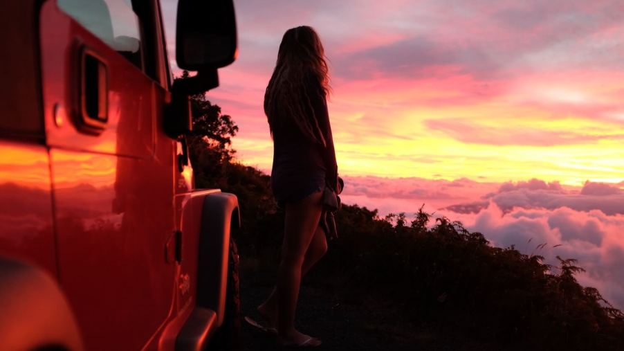 A woman stands next to a Jeep Wrangler, watching the sun set from atop a mountain.
