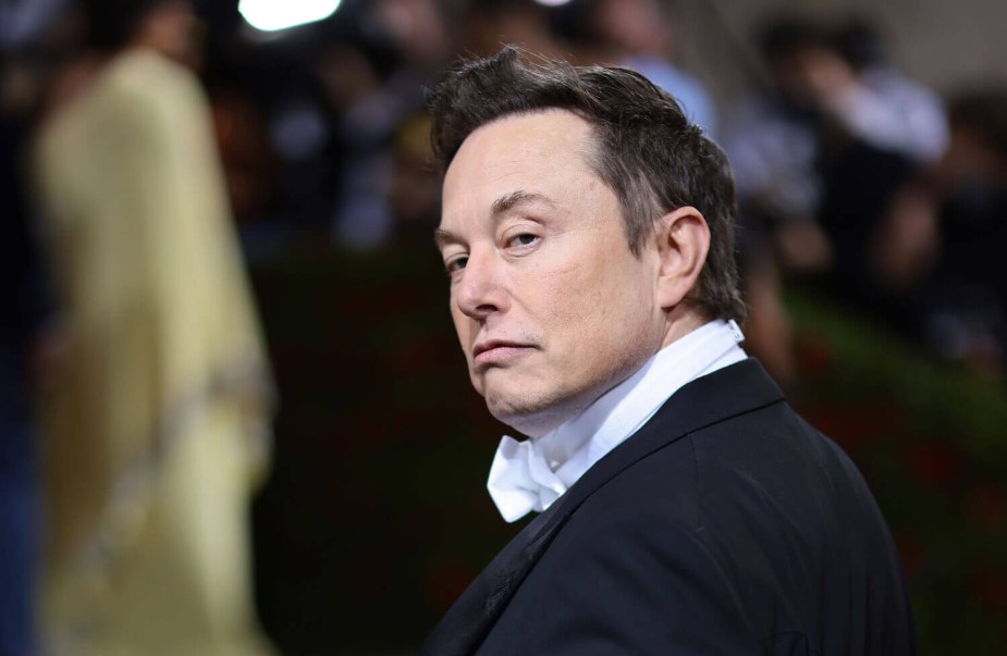 Elon Musk dressed in a Tuxedo, the met gala dinner behind him.