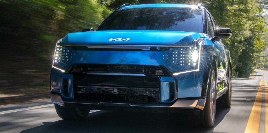A blue 2024 Kia EV9 midsize electric SUV is driving on the road. 