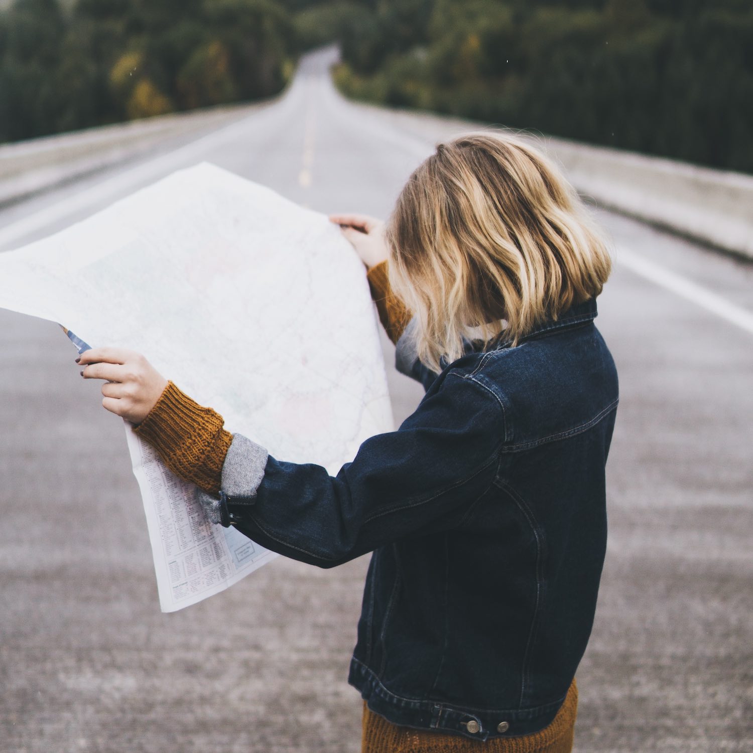 Blonde woman stands in the center of an interstate highway while examining an unfolded map.