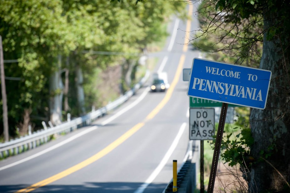 A blue "Welcome to Pennsylvania Sign" with a tree-lined two-lane road visible in the background.