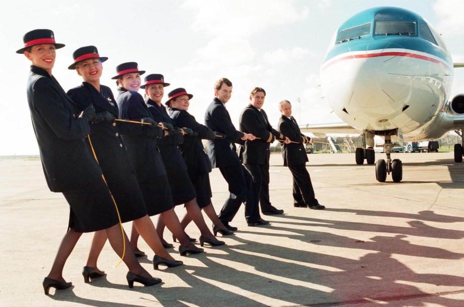 A group of smiling airline employees hold a rope and prepare to tow a jet parked in the background.