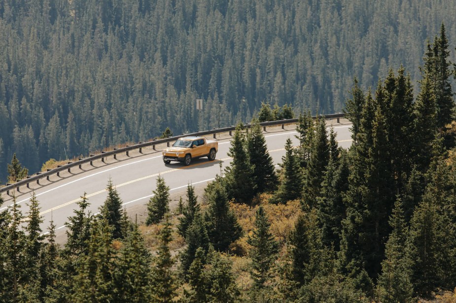 Overhead view of a yellow Rivian R1T electric truck cornering on a mountain road, pine trees visible in the background.