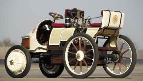 A white replica of the 1899 Porsche Semper Vivus EV parked on pavement, a fence in the background.