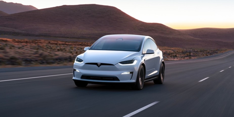 A white 2023 Tesla Model X midsize SUV is driving on the road.