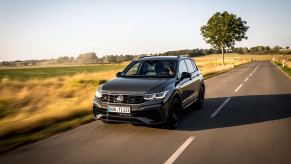 A gray 2023 Volkswagen Tiguan drives down a road with grass and a tree in the background. The Tiguan is the cheapest three-row SUV of 2023.