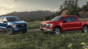 Blue and red versions of the 2022 Ford F-150, one of the least reliable 2022 pickup trucks, parked in a field of grass near a mountain range