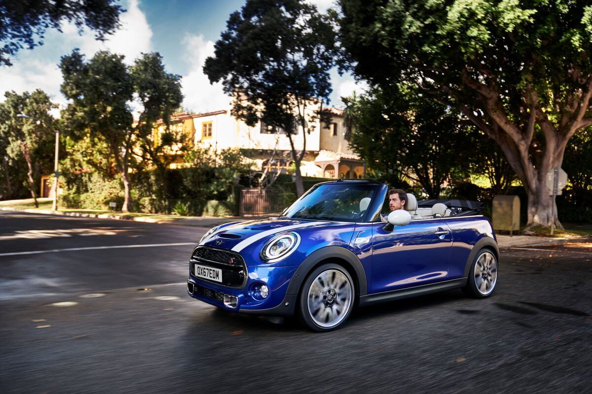 A blue and white 2019 Mini Convertible compact car driving through an intersection in a suburb