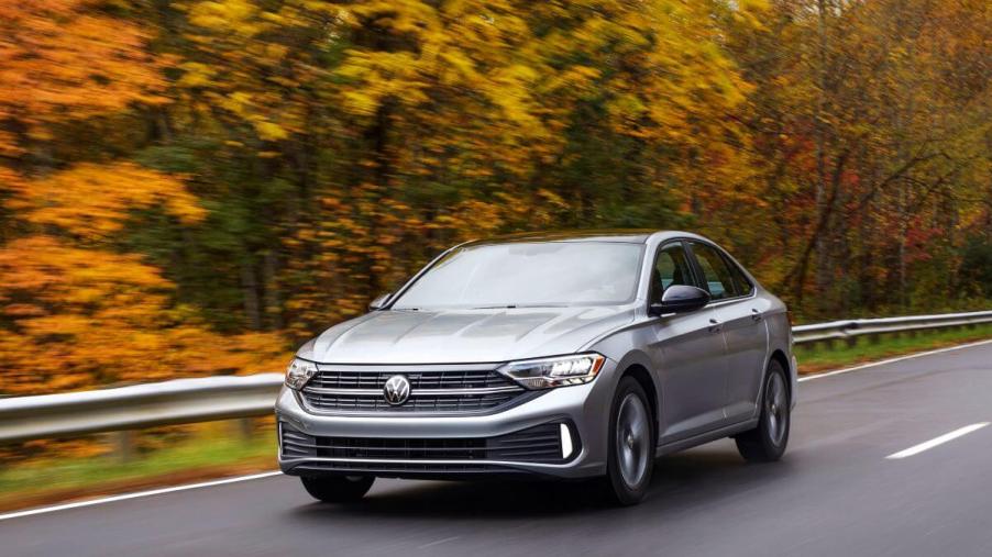 A silver-gray 2022 Volkswagen Jetta compact sedan model driving on a highway surrounded by a forest of yellow trees