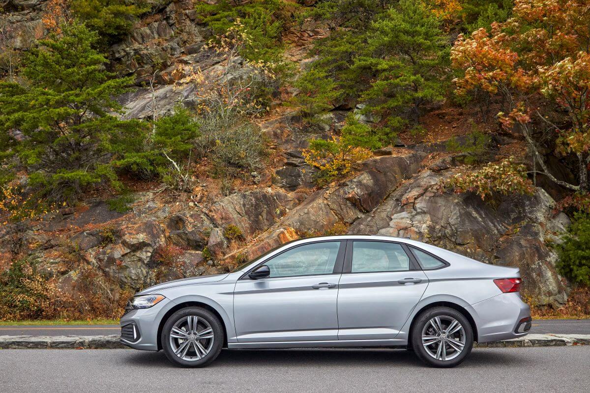 A side profile of a gray 2022 Volkswagen Jetta compact sedan model parked next to a wall of rocks and greenery