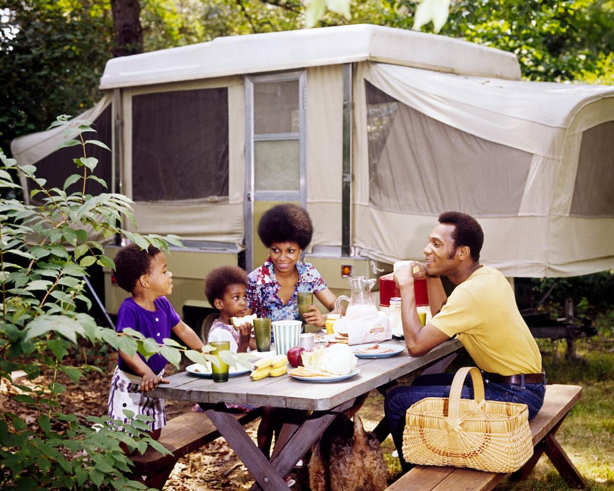 A smiling 1970s family eats at a picnic table in front of a pop-up camper