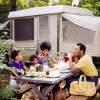 A smiling 1970s family eats at a picnic table in front of a pop-up camper