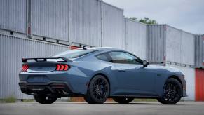 A rear exterior side shot of a blue-gray 2024 Ford Mustang muscle car coupe model parked near shipping containers. The 2024 Ford Mustang fuel economy gives it a big advantage over the competition.