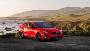 A red 2024 Subaru Impreza RS compact hatchback model parked on a grassy ledge near waves crashing in the sea